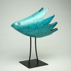 Op de afbeelding: Een turquoise vogelsculptuur met een craquelé glazuurafwerking, voorzien van een spitse snavel, één zichtbaar oog en vleugelachtige staartveren. De vogel staat op twee dunne, donkere metalen poten op een vierkante zwarte metalen voet.