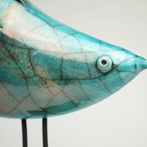 Op de afbeelding: Een keramieken vogelsculptuur met een glanzend turquoise glazuur en een opvallend donker craquelépatroon. De vogel heeft een spitse snavel, een rond oog en staat op twee dunne, donkere metalen poten met een glad, reflecterend oppervlak.