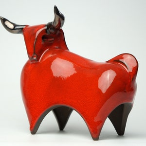 Puede incluir: Escultura de cerámica de un toro rojo con cuernos y patas negras. El toro tiene un acabado brillante y un diseño estilizado. La escultura es un objeto decorativo, probablemente para la decoración del hogar.