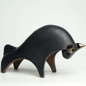 Op de afbeelding: Zwarte keramische stiersculptuur met bronzen hoorns en accenten. De stier heeft een gestileerd, modern ontwerp met een gebogen rug en poten. Op een witte achtergrond.