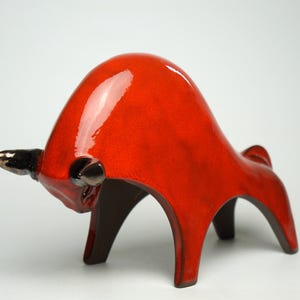 Könnte beinhalten: Eine Stierfigur aus Keramik mit einer glänzenden, leuchtend roten Glasur auf dem gewölbten Rücken und Körper. Der Stier hat dunkel metallisch wirkende Hörner und Ohren, eine gesenkte Kopfhaltung und vier kräftige Beine in Angriffsposition.