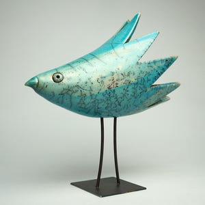 Op de afbeelding: Een turquoise keramieken vogelsculptuur met een craquelé glazuurafwerking en donkere, dunne, vertakte patronen. De vogel heeft een puntige, waaiervormige staart, één zichtbaar oog en staat op twee dunne, donkere metalen poten op een voet.