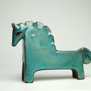 Può includere: Una statuetta di cavallo in ceramica stilizzata con una smaltatura turchese lucida e vibrante, con sfumature marrone scuro visibili lungo i bordi e la base. La scultura presenta una criniera fluida e una coda curva su quattro zampe.