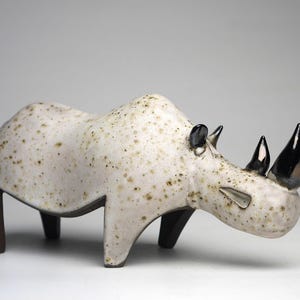 May include: A ceramic rhinoceros figurine featuring a speckled off-white glaze over a smooth body. The sculpture has two prominent, glossy black horns, small black ears, and dark brown legs that extend downwards from the main body structure.