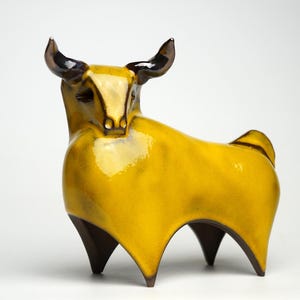 Può includere: Una statuetta di toro in ceramica gialla con corna e gambe nere. Il toro è in piedi su una superficie bianca.