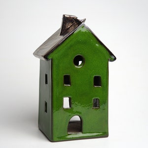 May include: A ceramic house-shaped candle holder with a glossy green finish. The house has a dark brown roof and chimney, with cut-out windows and a round opening. The design is a miniature house, perfect for home decor.