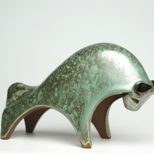 Peut inclure: Une sculpture de taureau en céramique verte avec un glaçage moucheté et des cornes en bronze. Le taureau est debout sur quatre pattes et a un corps incurvé.