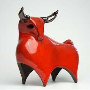 Può includere: Scultura in ceramica di un toro rosso acceso. Le corna e le zampe sono marroni scuro. L'oggetto decorativo, con finitura lucida, è destinato all'arredamento di una casa o di un ufficio.