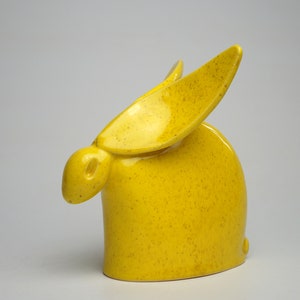 Op de afbeelding: Een gele keramische konijnenfiguur met een gespikkelde textuur. Het konijn heeft lange, rechtopstaande oren en een afgerond lichaam. Het beeldje is een decoratief item, waarschijnlijk voor woondecoratie.