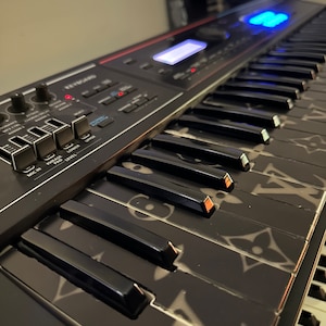 May include: A black keyboard with a custom Louis Vuitton design. The keyboard has a digital display and several knobs and buttons.