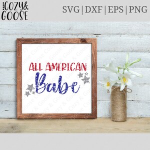 May include: A framed sign with a distressed red, white, and blue graphic that reads "All American Babe" with stars around the text. The sign is on a wooden surface with white flowers in a vase.