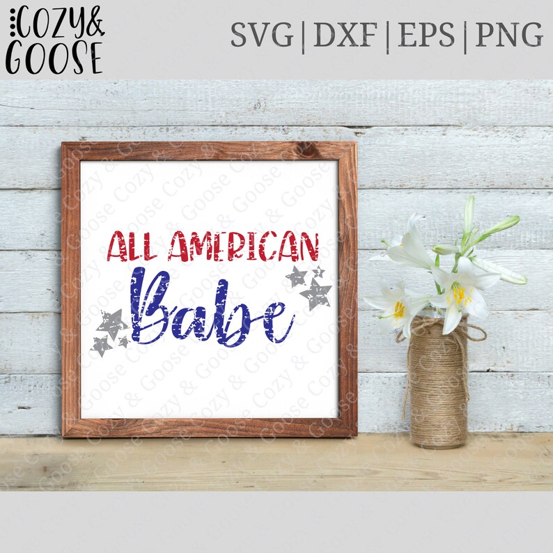 May include: A framed sign with a distressed red, white, and blue graphic that reads "All American Babe" with stars around the text. The sign is on a wooden surface with white flowers in a vase.