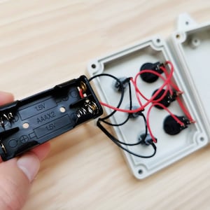 May include: A black battery pack with two AAA batteries and the text "1.5V AAAx2 1.5V" printed on it. The battery pack is connected to a white plastic box with wires and three black round components.