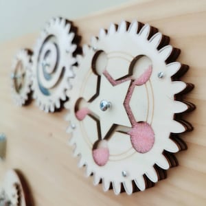May include: Close-up of a wooden toy with three interlocking gears. The largest gear has a pink and white design and is made of layered wood. The gears are mounted on a wooden surface.