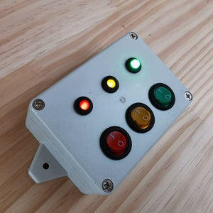 May include: A white plastic box with four buttons and three lights. The buttons are black with a red, yellow, and green light inside. The lights are red, yellow, and green.