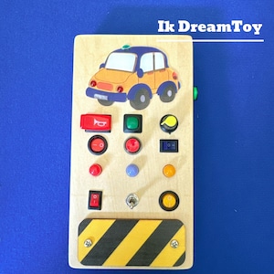 May include: A wooden toy panel with a yellow car, various colored buttons, and a black and yellow striped panel at the bottom. The panel is labeled "Ik DreamToy".
