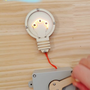 Könnte beinhalten: Ein Holzspielzeug in Form einer Glühbirne mit einem roten Kabel, das mit einer grauen Box mit einem Holzhebel verbunden ist. Die Glühbirne hat ein glühendes Filament im Inneren.