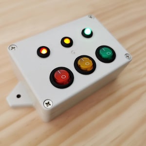 Può includere: Una scatola bianca con quattro interruttori a levetta e quattro spie luminose. Le luci sono rossa, gialla, verde e bianca. Gli interruttori sono neri con pulsanti rosso, giallo, verde e bianco.