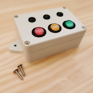 May include: A white plastic box with four toggle switches. The switches are red, yellow, green, and black. Two silver screws are visible on the wooden surface.