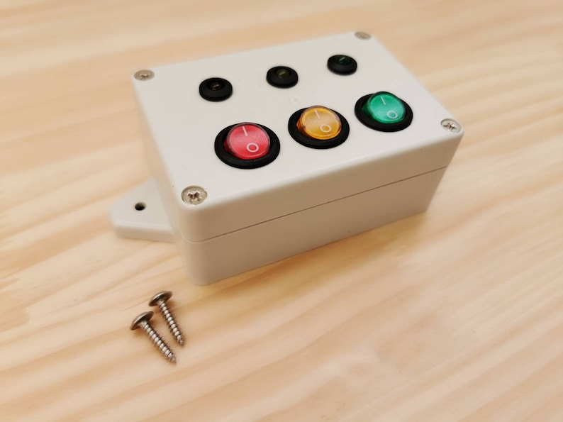 May include: A white plastic box with four toggle switches. The switches are red, yellow, green, and black. Two silver screws are visible on the wooden surface.