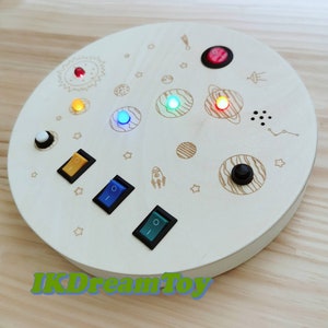 Puede incluir: Un juguete redondo de madera con temática espacial. Presenta un sol, planetas, estrellas, un cohete y un OVNI. Hay cinco botones de diferentes colores: rojo, naranja, amarillo, azul y verde. También hay tres interruptores de palanca y un botón negro. El juguete tiene el texto "IKDreamToy" impreso en la parte inferior.