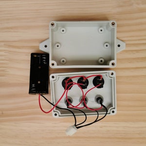 May include: A white plastic box with three black electrical components and red and black wires. The box is open and the components are visible. A black battery pack is connected to the box with wires.
