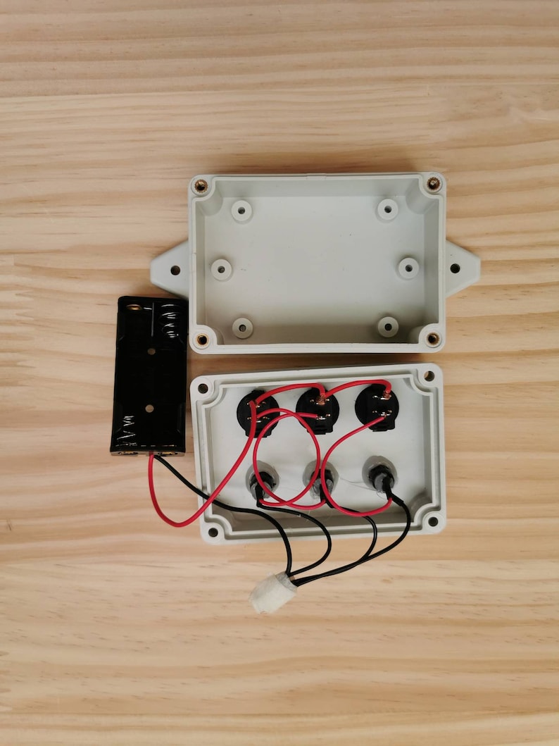May include: A white plastic box with three black electrical components and red and black wires. The box is open and the components are visible. A black battery pack is connected to the box with wires.
