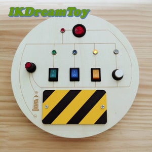 May include: A circular wooden sensory board featuring the text IKDreamToy, a large red power switch, four small indicator lights in red, green, blue, and yellow, three rectangular toggle switches in green, blue, and orange, and a yellow and black striped plate.