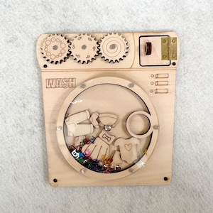 May include: A light-coloured wooden sensory board shaped like a washing machine with the word WASH engraved on the front. It features three rotating gears, a hinged door, and a circular window containing small wooden clothing shapes and colourful beads.