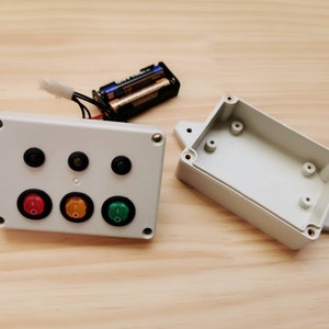A white plastic project box with three toggle switches and three LEDs. The box is open and the lid is next to it. Two AA batteries are visible in the background.