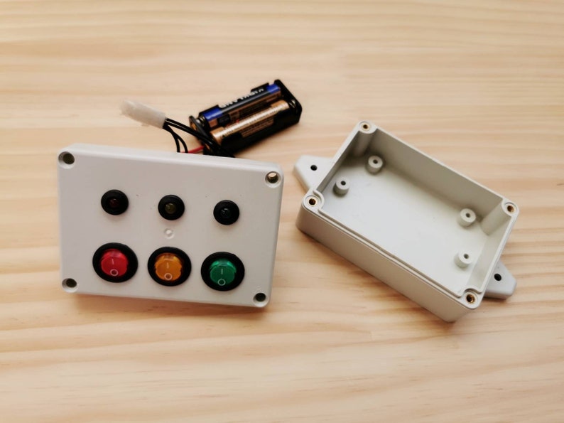 A white plastic project box with three toggle switches and three LEDs. The box is open and the lid is next to it. Two AA batteries are visible in the background.