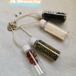 May include: Four small clear glass bottles with white lids, each filled with different types of small seeds. The bottles are attached to a white string with a metal clasp. The bottles are on a white surface. The text "IK DreamToy" is visible in the top left corner of the image.