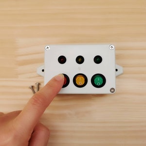 May include: A white plastic box with three buttons and three indicator lights. The buttons are red, yellow, and green. The lights are red, yellow, and green. The box is mounted on a wooden surface.