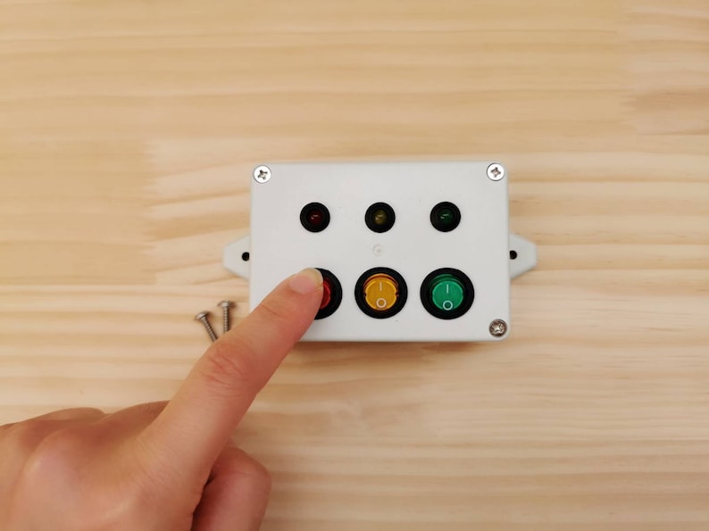 May include: A white plastic box with three buttons and three indicator lights. The buttons are red, yellow, and green. The lights are red, yellow, and green. The box is mounted on a wooden surface.