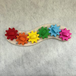 May include: A wooden toy with six brightly coloured gears: red, orange, yellow, green, blue, and pink. The gears are mounted on a curved, light-coloured wooden base. Each gear has a matching coloured handle.
