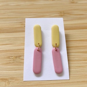 Puede incluir: Un par de pendientes colgantes de color amarillo y rosa. La parte superior de los pendientes es amarilla y la parte inferior es rosa.