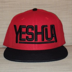 May include: A red and black baseball cap with the word "YESHUA" embroidered in black on the front.