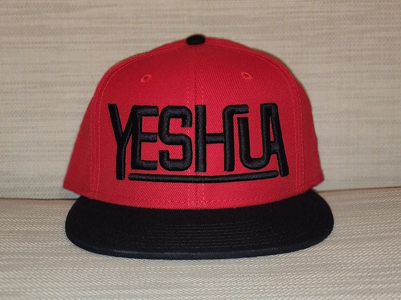 May include: A red and black baseball cap with the word "YESHUA" embroidered in black on the front.