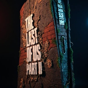 Può includere: Una scultura dettagliata di una struttura in pietra erosa con la scritta "THE LAST OF US PART II" in bianco. La scultura ha una texture di mattoni e pietra, con muschio verde e piante.
