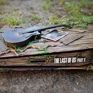 May include: A custom PlayStation 5 console with a wooden design, a guitar, a photo, and wooden slats. The console is decorated with green moss and branches, and the text "THE LAST OF US PART II" is visible.