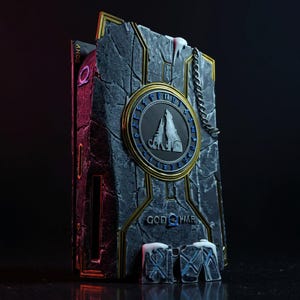 May include: A stone-textured God of War collectible. The front features a circular emblem with a mountain and wolf design, surrounded by gold and blue accents. The words "GOD OF WAR" are visible. A chain and small stone blocks complete the design.