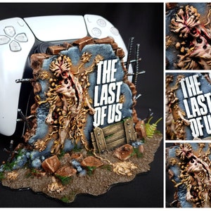 May include: A white PlayStation 5 controller sits on a detailed stand. The stand has a weathered stone design with the text "THE LAST OF US" and a zombie-like figure, in a brown and grey colour scheme.