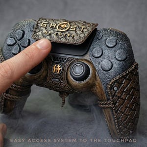 Peut inclure: Une manette de jeu vidéo personnalisée avec une finition texturée marron et noire. La manette présente le texte "GHOST OF TSUSHIMA" en lettres dorées et un symbole japonais. Le design comprend un motif texturé unique.