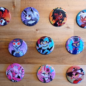 Op de afbeelding: Een verzameling van tien ronde buttons met kleurrijke cartoonkarakterontwerpen. De buttons tonen verschillende karakters met levendige kleuren, waaronder rood, paars, blauw en roze, tegen een houten achtergrond.