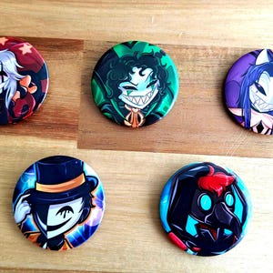 May include: Five circular pin-back badges featuring stylised character illustrations. The top row shows a white-haired figure in red, a dark-haired figure in green, and a blue-haired figure in purple. The bottom row shows a masked figure in a top hat and a dark hooded figure with a beak mask.