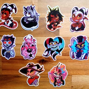 May include: A collection of colorful cartoon character stickers. The stickers feature various demons and imps with unique designs, including horns, tails, and expressive faces. The stickers are on a wooden surface.