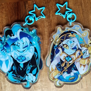 May include: Two acrylic keyrings featuring cartoon characters. One is blue and black with a crown and a sinister grin. The other is white, blue, and gold, holding a staff. Each keyring has a blue star-shaped clasp.