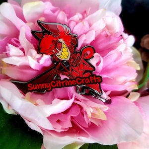 Könnte beinhalten: Ein rot-schwarzer Emaille-Pin mit einer Cartoon-Figur mit breitem Grinsen und spitzen Zähnen. Der Pin ist vor einem Hintergrund aus rosa Blüten platziert. Der Text "SunnyCitrineCrafts" ist auf dem Pin zu sehen.