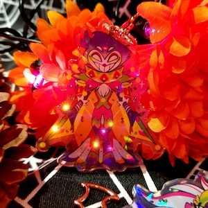 Könnte beinhalten: Ein holografischer Anhänger mit einer Comicfigur in einem lila und schwarzen Outfit, einem gelben Stern und einem rot-weiß gestreiften Kragen. Die Figur hat ein breites Lächeln und eine rote Nase. Der Anhänger ist von einem glitzernden, schillernden Hintergrund umgeben.
