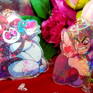 May include: Two iridescent holographic keychains featuring cartoon characters. One keychain is a red and white jester with a heart-shaped halo. The other keychain is a purple and blue angel with a heart-shaped halo. Both keychains have a holographic background with hearts.